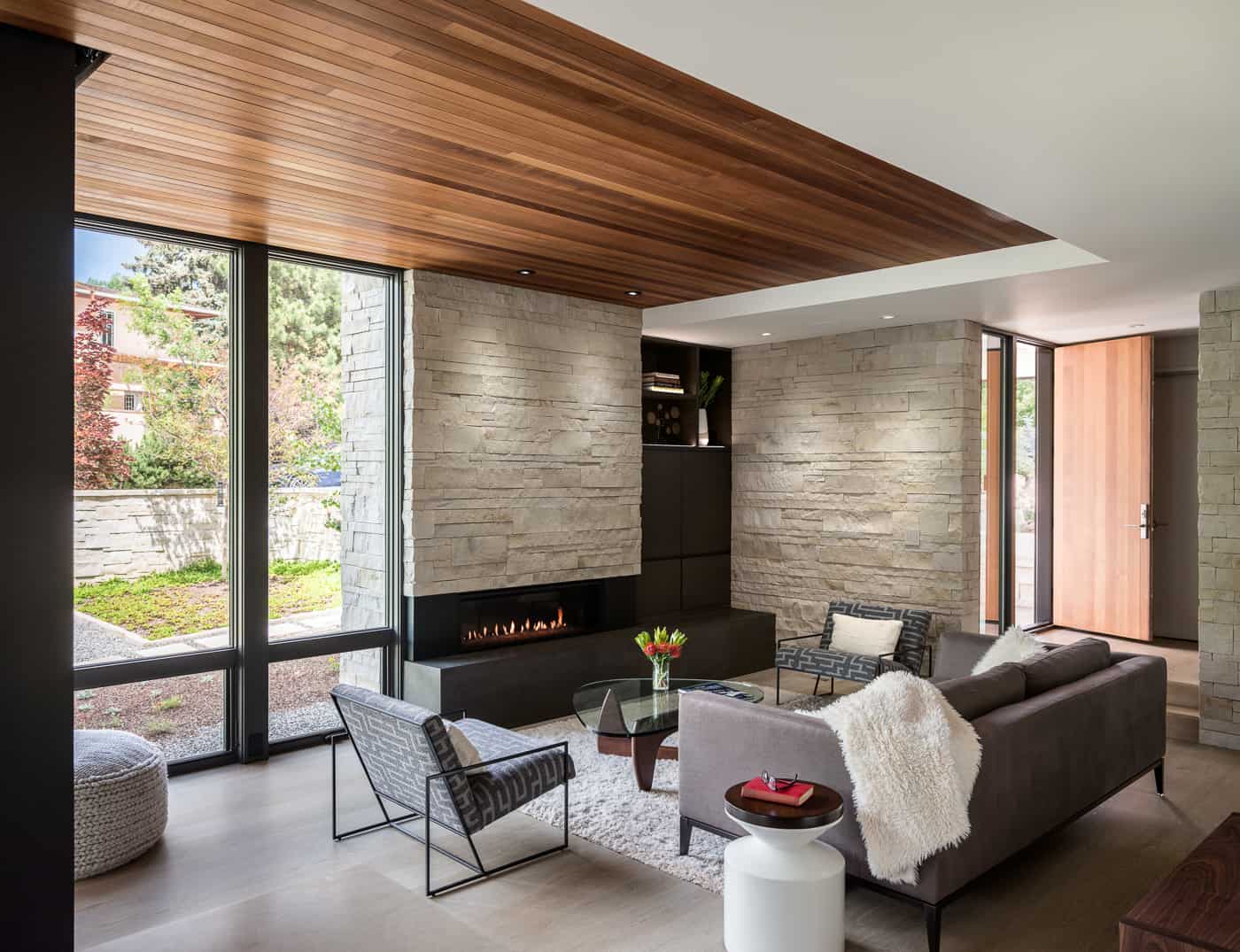 Minimalist modern living room with wood ceiling, stone fireplace, and floor to ceiling windows